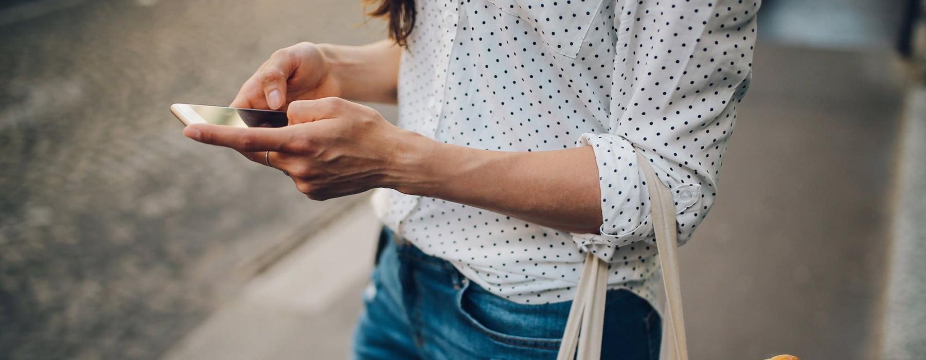 woman walks down the street and texts with a bag of groceries on her arm